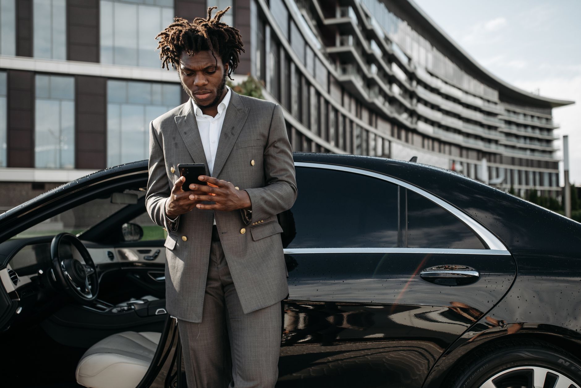 Homem de terno consultando o celular encostado em um carro